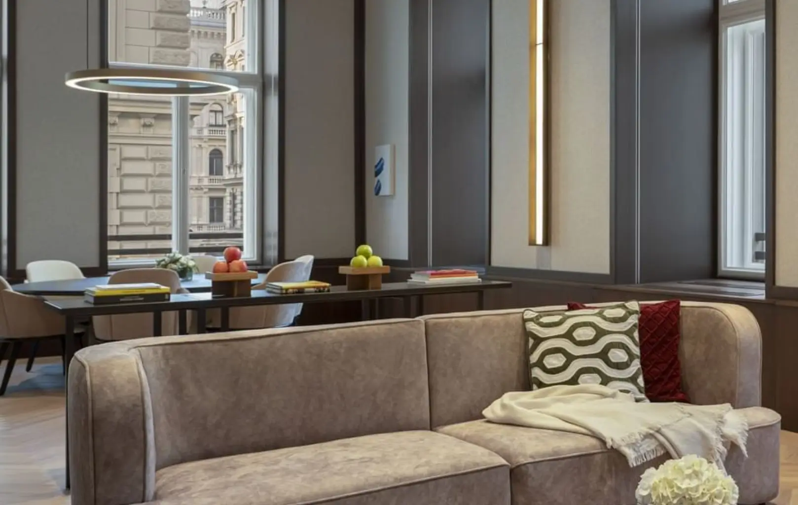 Ein modernes Wohnzimmer mit einem beigen Sofa, gemusterten Kissen, einer Decke und einem Esstisch mit Obstschalen in der Nähe der großen Fenster mit Blick auf die Gebäude der Stadt - ein echtes Suite-Erlebnis.